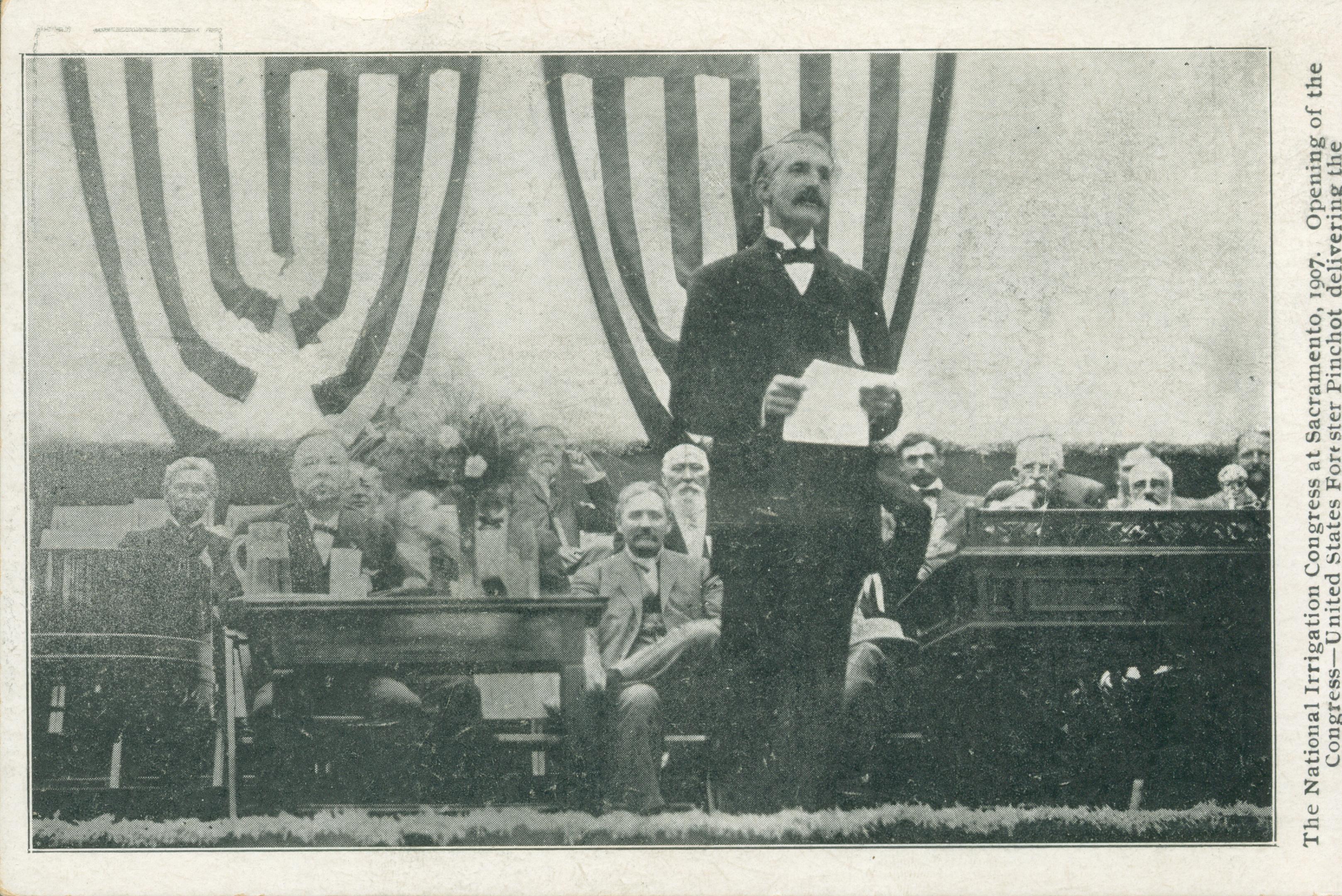 This postcard shows a Mr. Pinchot giving a speech with his back to several men seated at desks.